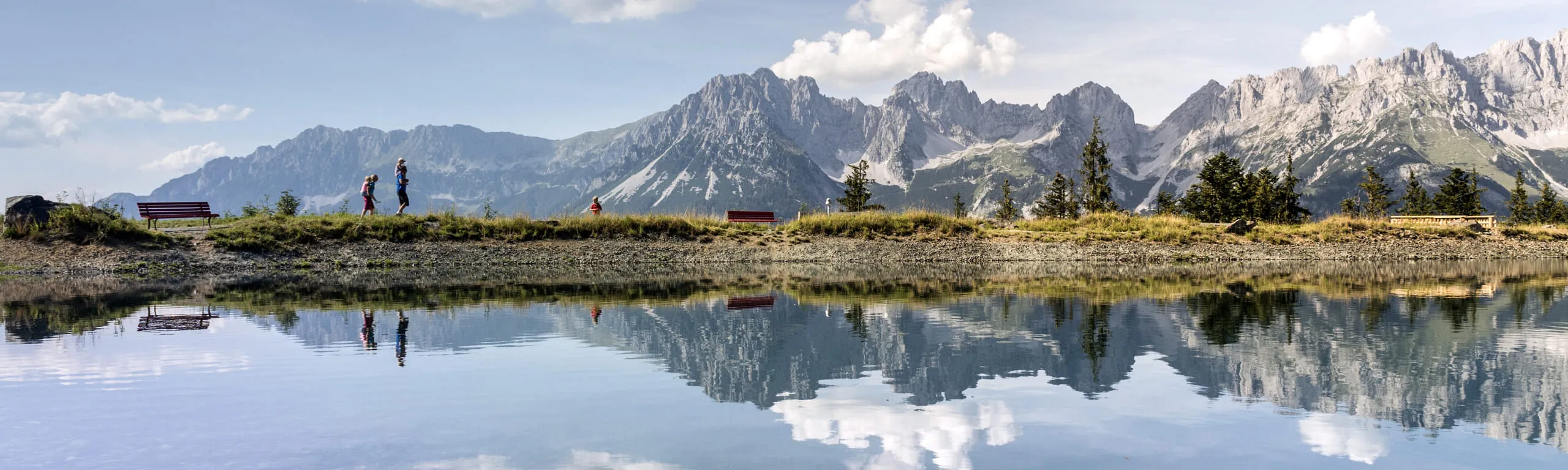 Panoramablick auf den Astbergsee mit Bergen im Hintergrund und Wanderern am Ufer.